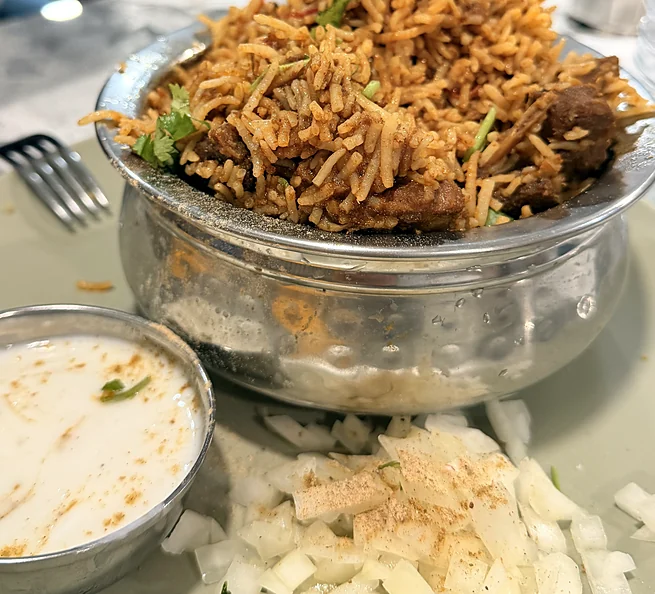 Biryani in a steel bowl with raita and chopped onion